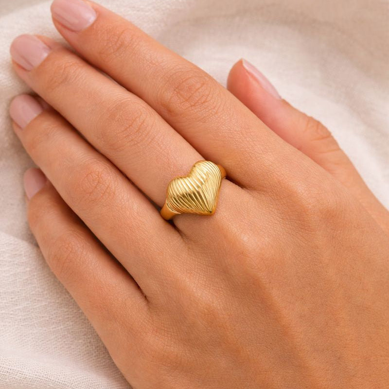 Gold heart-shaped ring on a hand against a beige background