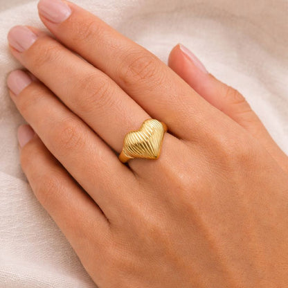 Gold heart-shaped ring on a hand against a beige background