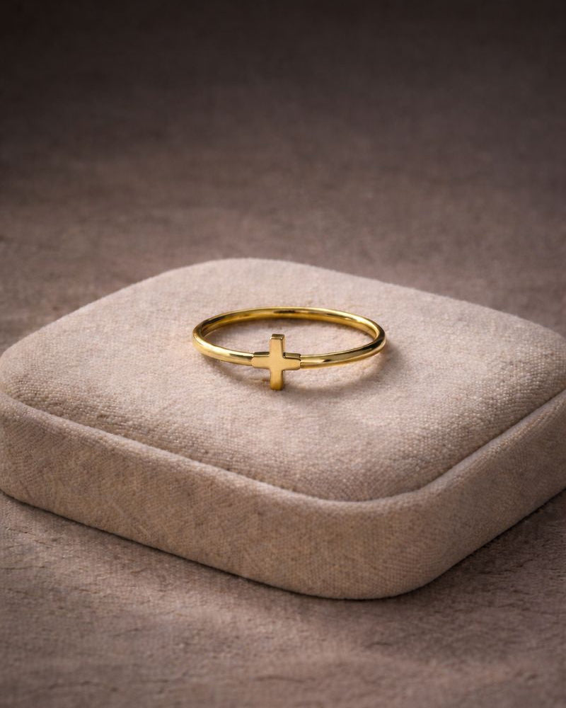 Gold ring with a cross design on a beige jewelry cushion against a dark background